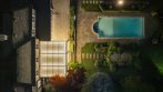 Aerial night view of an illuminated garden pergola beside a landscaped backyard pool.