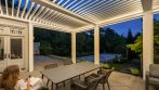 Outdoor dining area under open pergola louvers with integrated lighting at dusk.