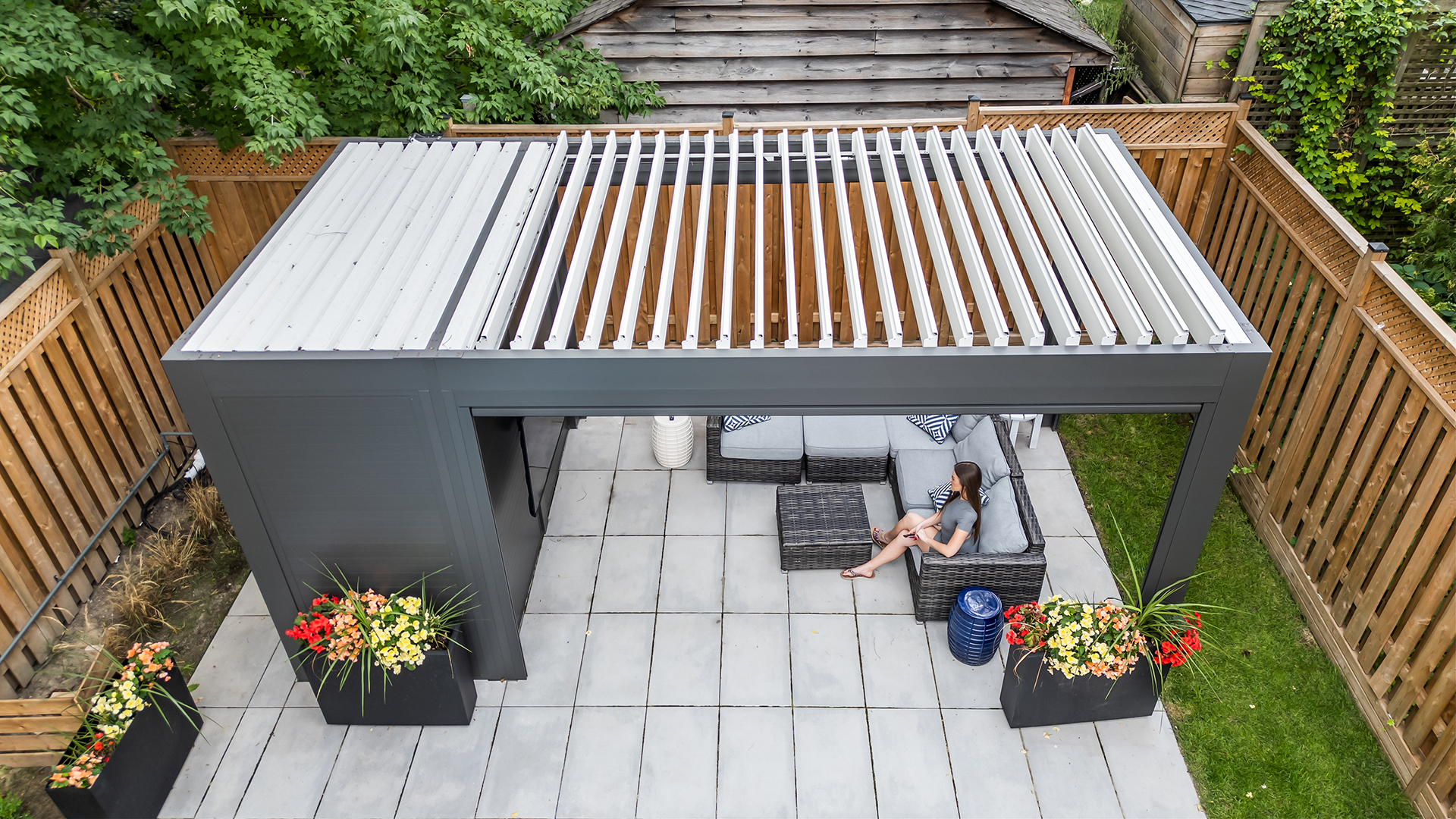 Aerial view of a shed pergola with white adjustable louvers open above a patio lounge area.