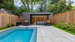 Shed with pergola at the end of a backyard swimming pool, framed by a wood fence and flower gardens.
