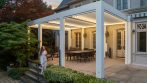 White louvered pergola with integrated lighting over a backyard patio dining space.