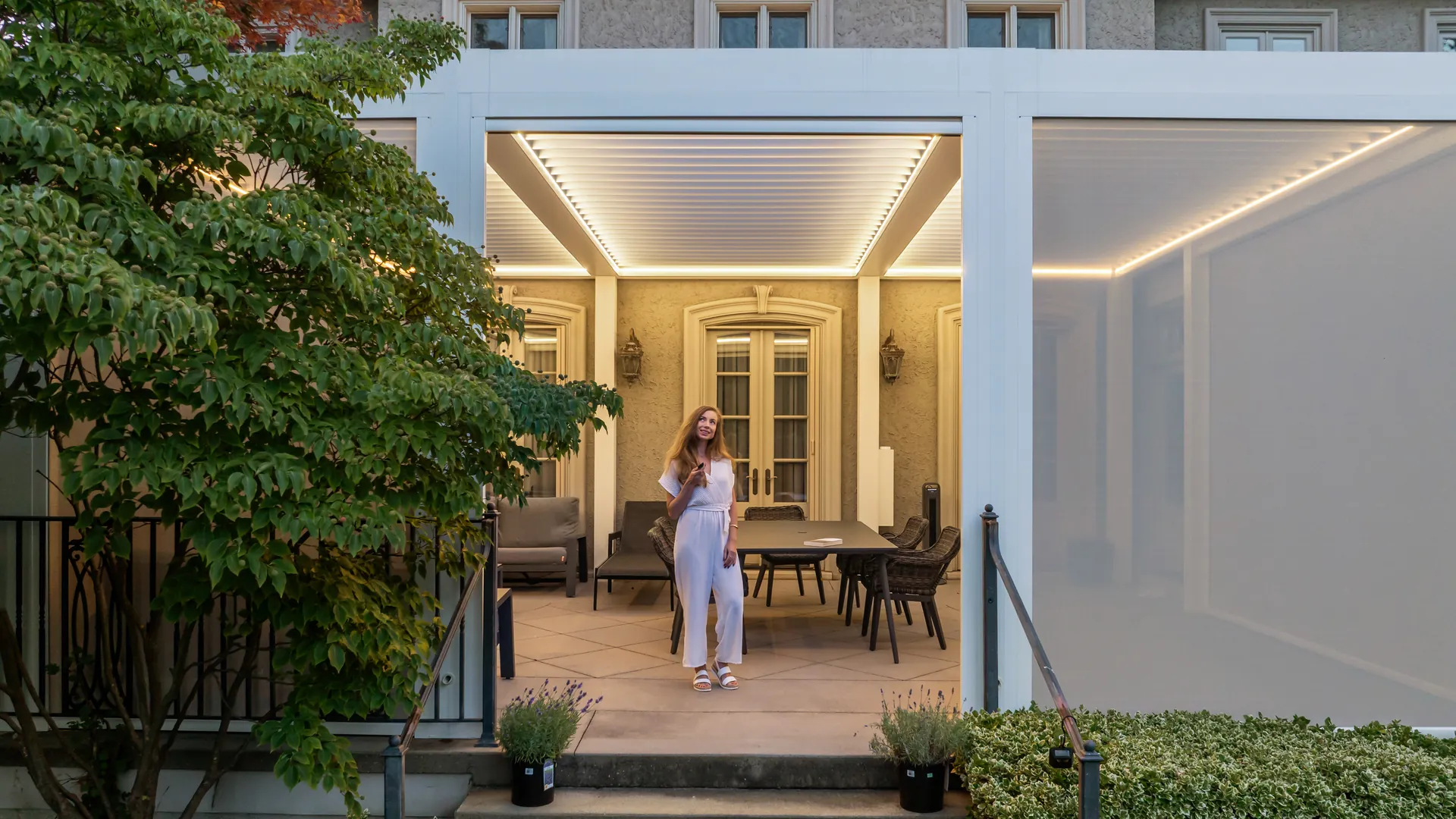 White attached louvered pergola with LED lighting and a lowered ZIP screen over a dining terrace by the garden steps
