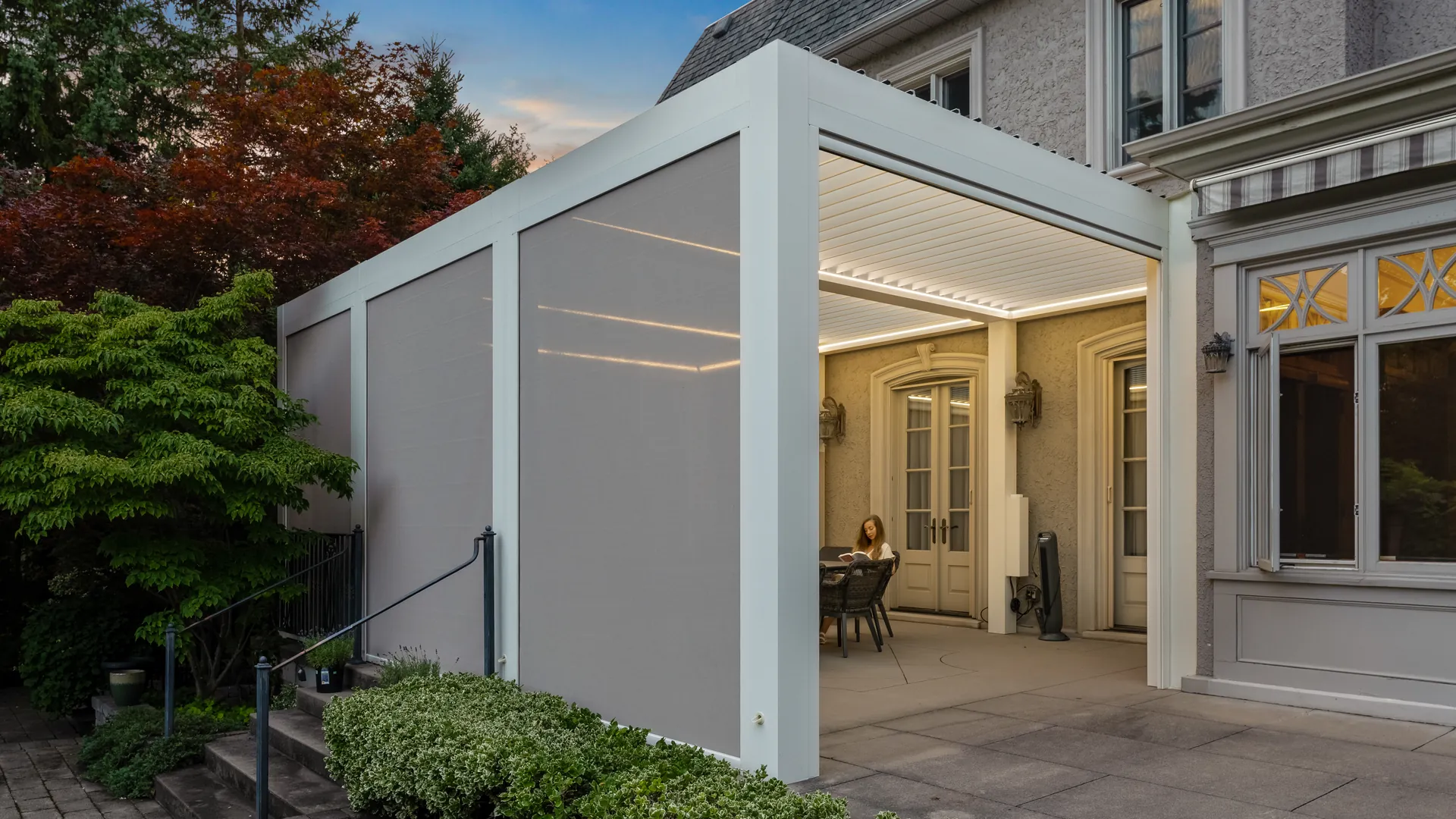 Pergola blanche adossée à lames orientables avec trois toiles de pergola beiges baissées sur un côté et éclairage DEL sur une table