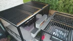 Backyard patio pergola with closed black louvers over a dining table, next to a stone fireplace and a hot tub pergola