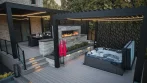 Black aluminum backyard pergola over a hot tub at dusk, with a stone fireplace and a second pergola over the dining area behind it