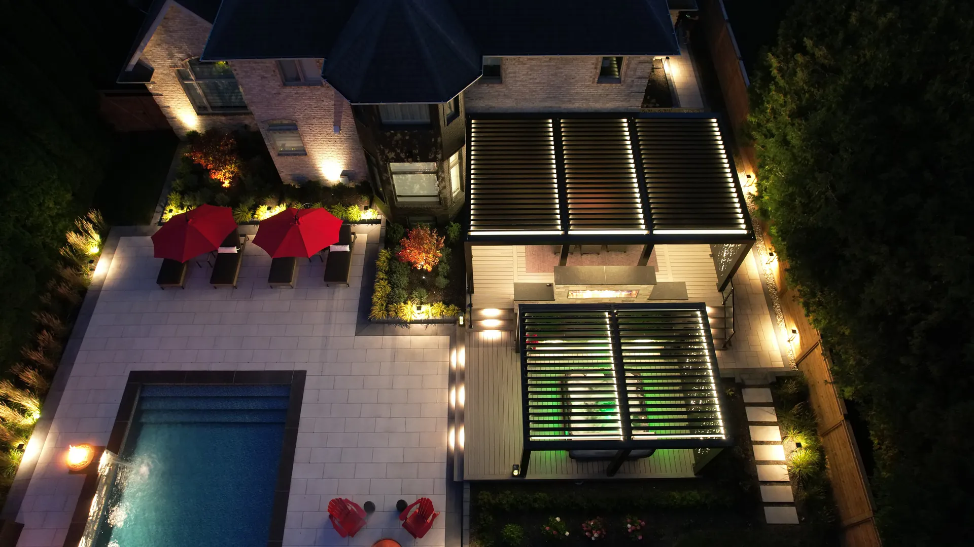 Night aerial view of two backyard pergolas with LED lights on, beside the pool patio and garden lighting