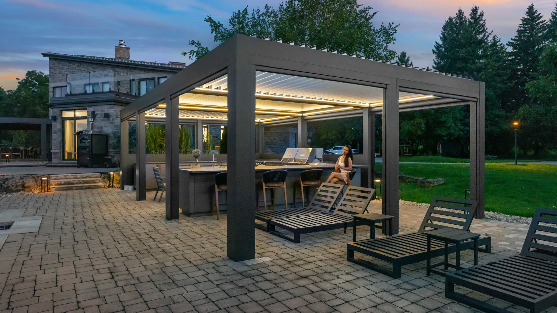 Custom aluminum louvered pergola with LED lighting over an outdoor kitchen and bar seating on a paver patio at dusk
