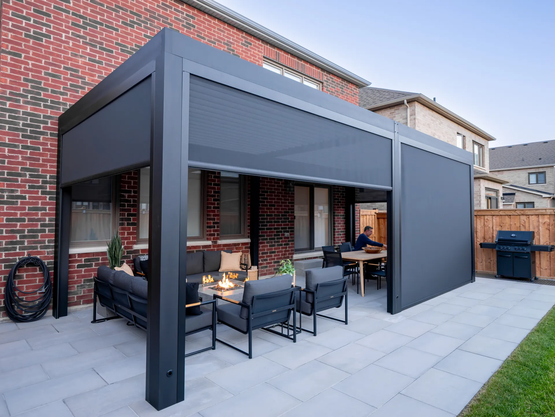 Pergola adossée en aluminium avec trois écrans ZIP à des hauteurs différentes autour d’un coin salon avec foyer et d’un coin repas