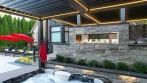 Black aluminum pergola with fireplace and grill seen from the hot tub, with the pool patio and red umbrellas on the left