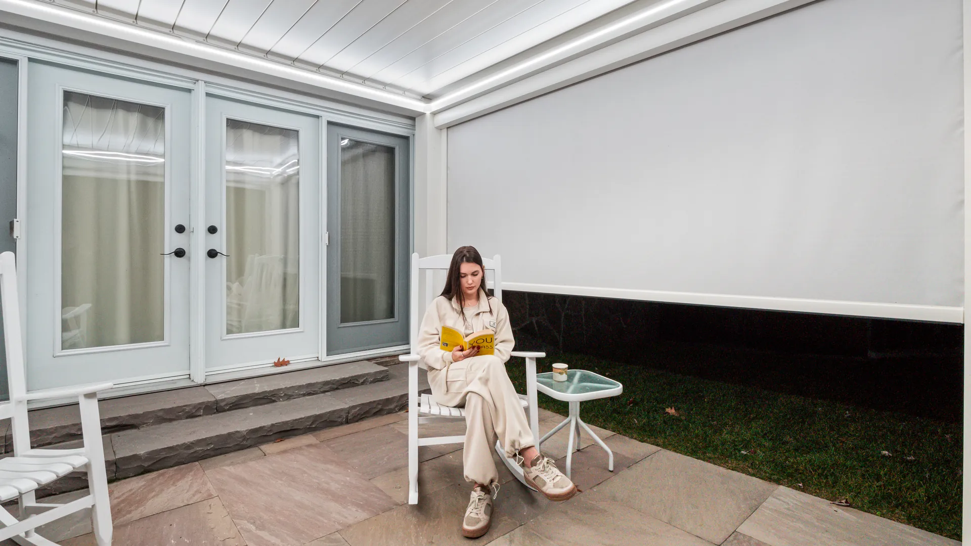 Woman reading under a white louvered pergola with a retractable pergola screen partly lowered on the yard side